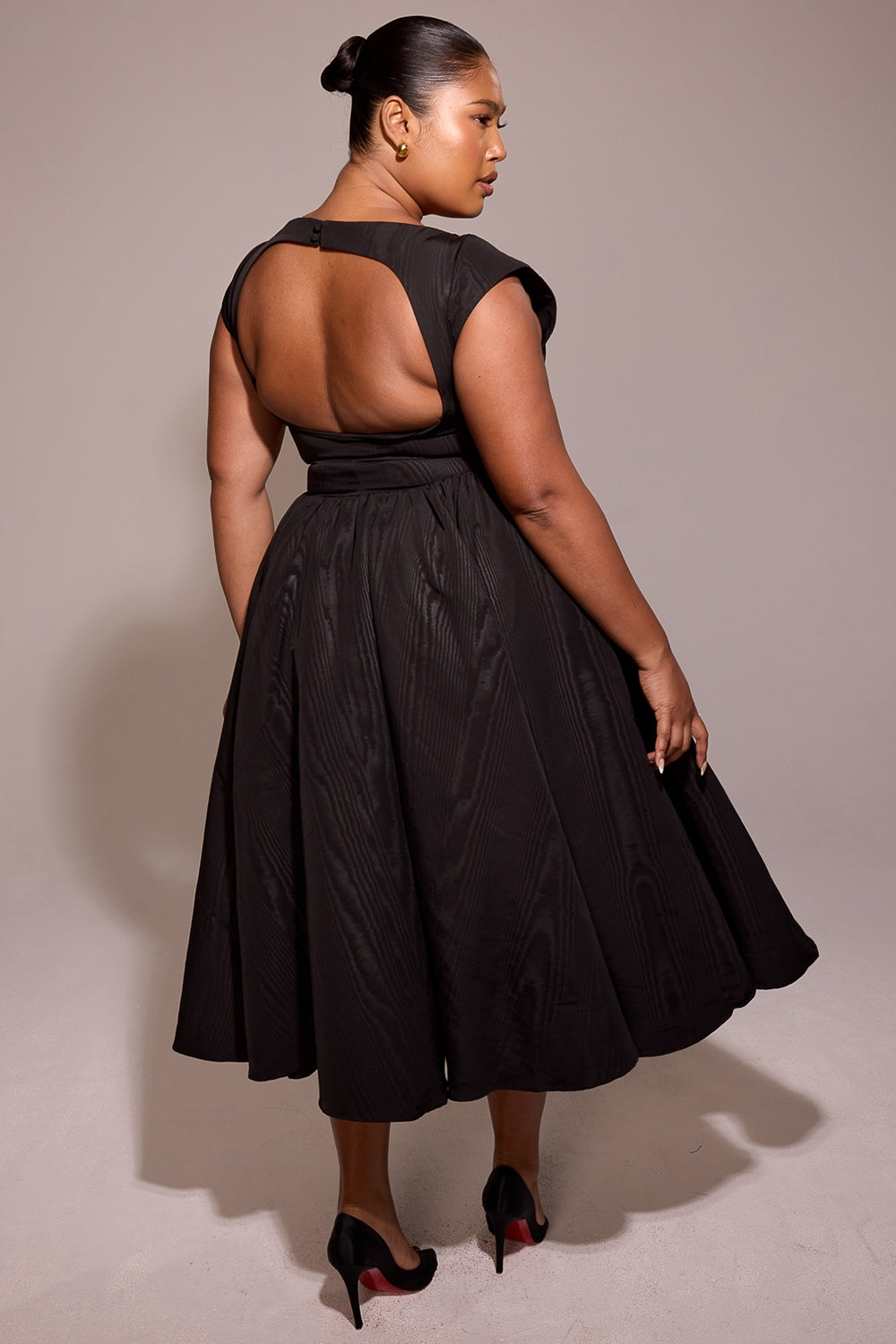 Back three-quarter view of black midi dress with scoop back cutout and wide skirt, warm studio backdrop