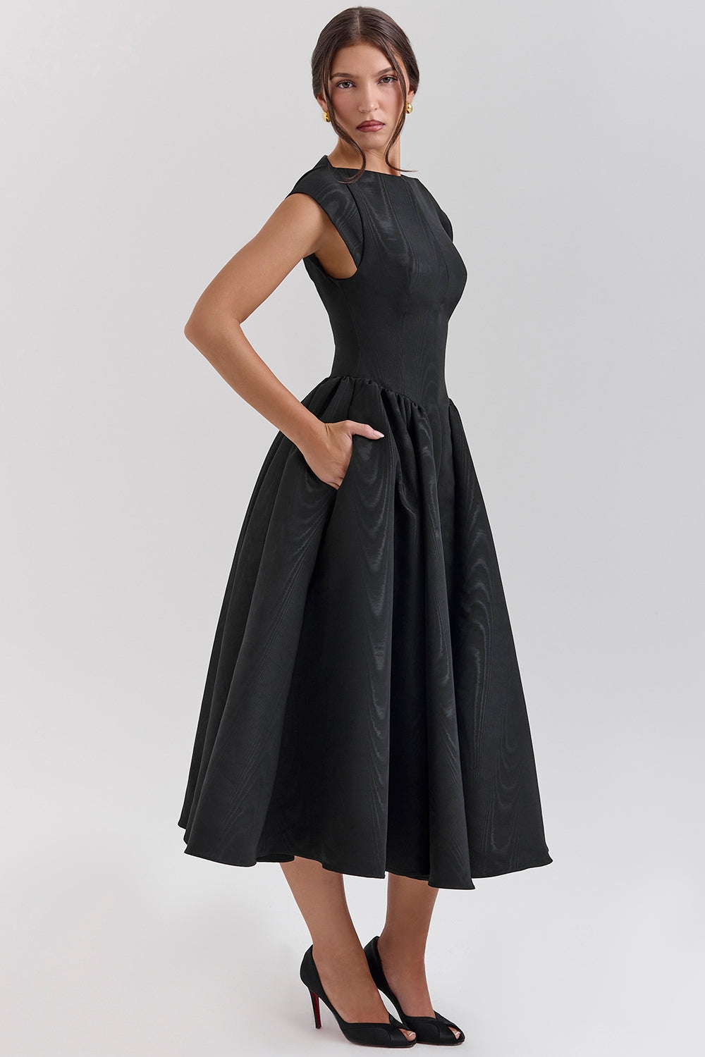 Black midi dress angled side view with hand in pocket, fitted bodice and flared skirt, satin sheen